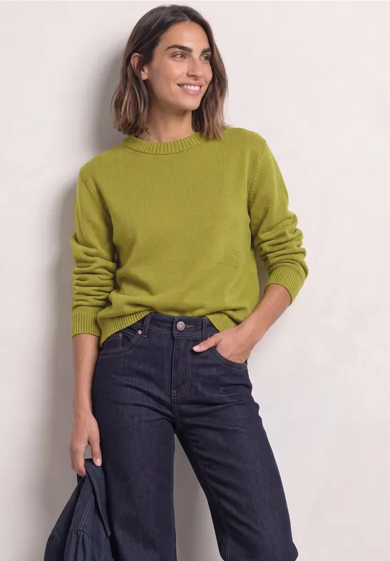 Eine Frau trägt einen grünen Pullover und dunkelblaue Jeans, hält eine Jacke in der Hand.