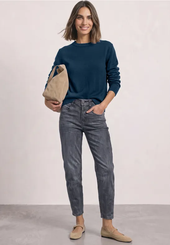 Frau in dunkelblauem Pullover, grauen Jeans und beigen Slippern hält eine beige Tasche vor neutralem Hintergrund.