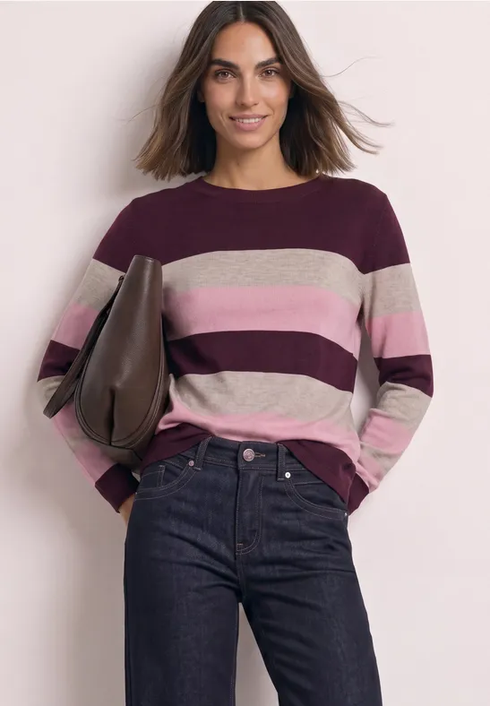 Eine Frau trägt einen gestreiften Pullover in Burgund-, Rosa- und Beigetönen und Jeans vor hellem Hintergrund.
