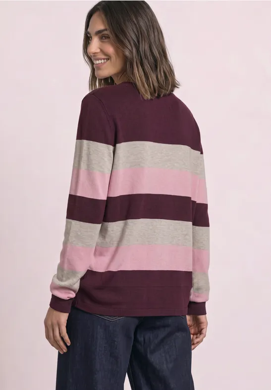 Eine Frau trägt einen breit gestreiften Pullover in Bordeaux, Rosa und Beige, schaut lächelnd über die Schulter.