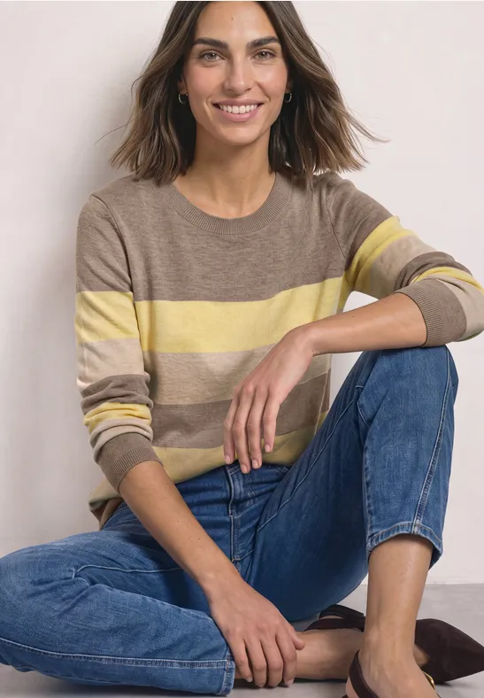 Eine lächelnde Frau in gestreiftem Pullover und Jeans sitzt entspannt auf dem Boden.