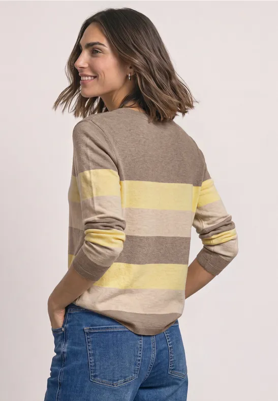 Frau mit schulterlangem Haar im gelb-beige gestreiften Pullover und Jeans schaut lächelnd über die Schulter.