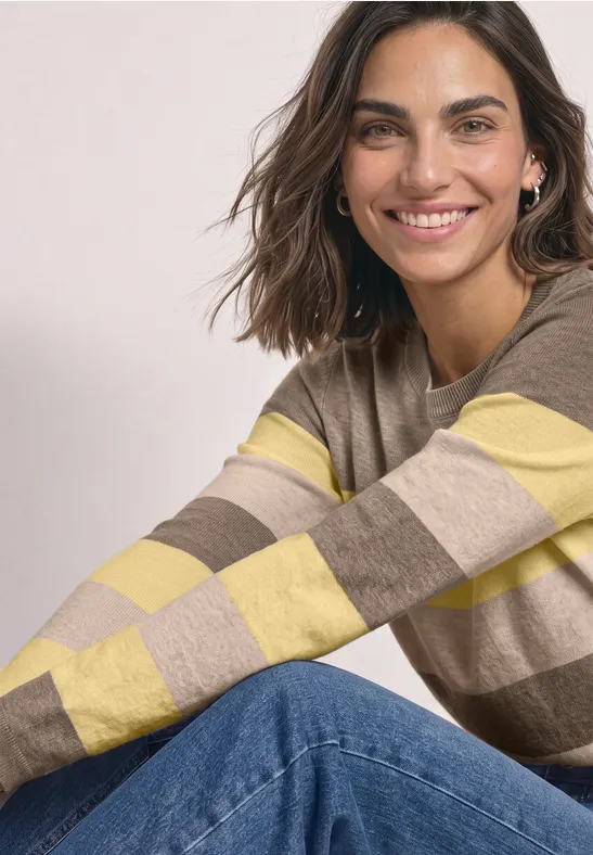 Eine lächelnde Frau trägt einen gestreiften beige-gelben Pullover und Jeans und sitzt entspannt.