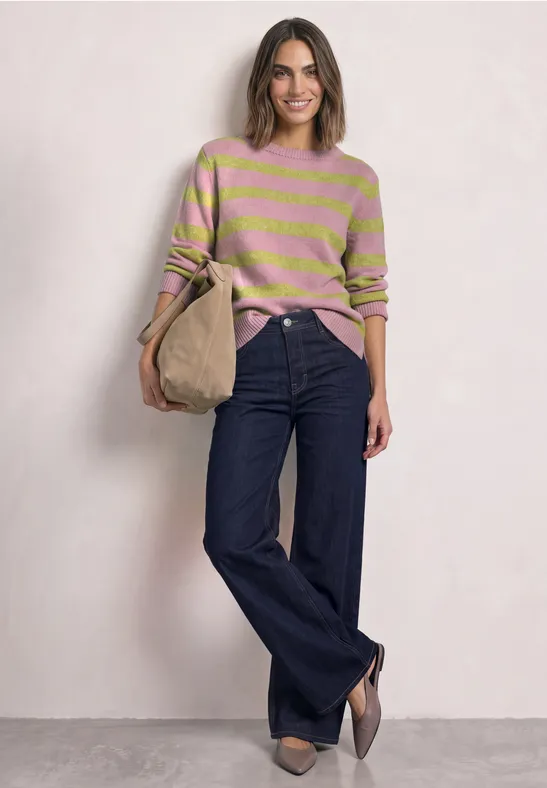 Frau in rosa-grün gestreiftem Pullover, weiten Jeans und Beuteltasche vor hellem Hintergrund.