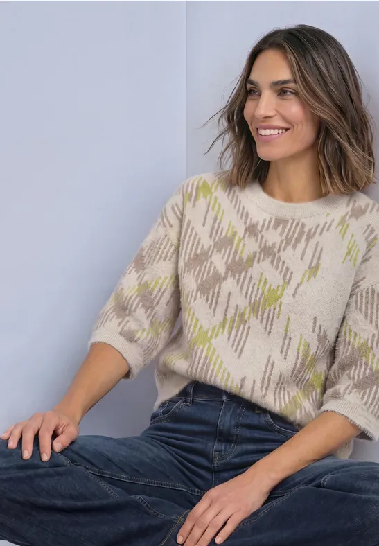 Eine lächelnde Frau sitzt entspannt, trägt einen kurzärmligen, gemusterten Pullover und Jeans.