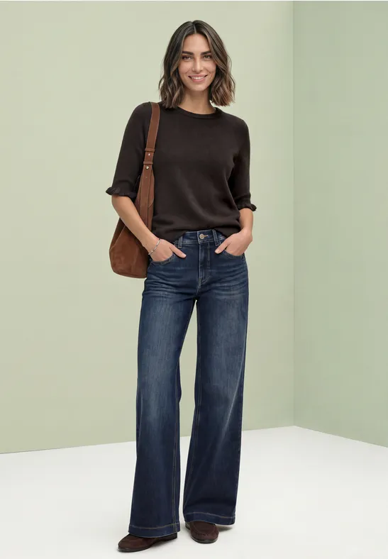 Frau mit braunem Kurzarm-Pullover, weitem Jeans und brauner Schultertasche vor hellem Hintergrund.