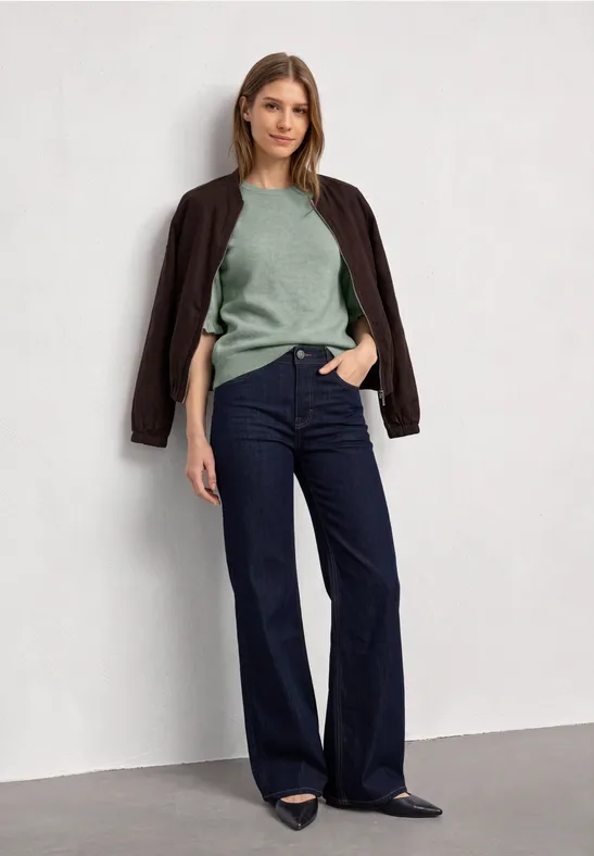 Eine Frau steht vor weißer Wand, trägt grünen Pullover, Jacke über den Schultern und weite Jeans.