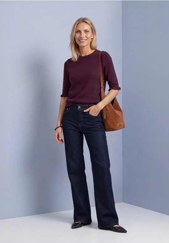 Eine blonde Frau in bordeauxfarbenem Pullover, weiten Jeans und brauner Tasche vor neutralem Hintergrund.