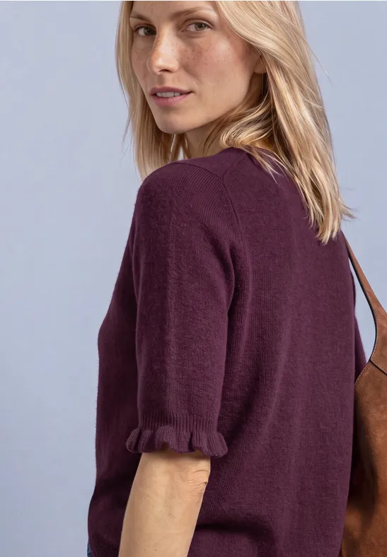 Blonde Frau im Dreiviertelprofil mit lila Shirt mit Rüschenärmeln und brauner Tasche.