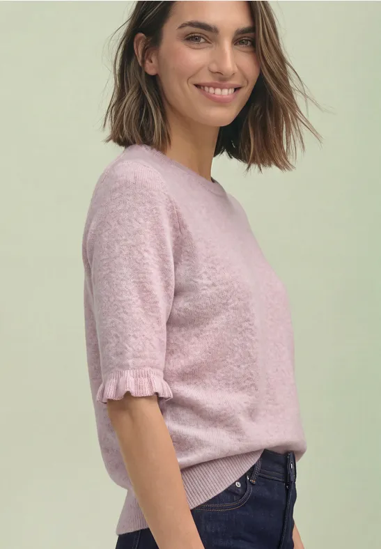 Frau in rosa Kurzarm-Pullover mit Rüschenärmeln, lächelnd seitlich posierend