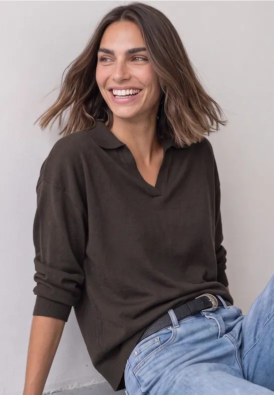 Une femme souriante aux cheveux châtains mi-longs, en pull marron foncé et jean, assise devant un mur neutre.