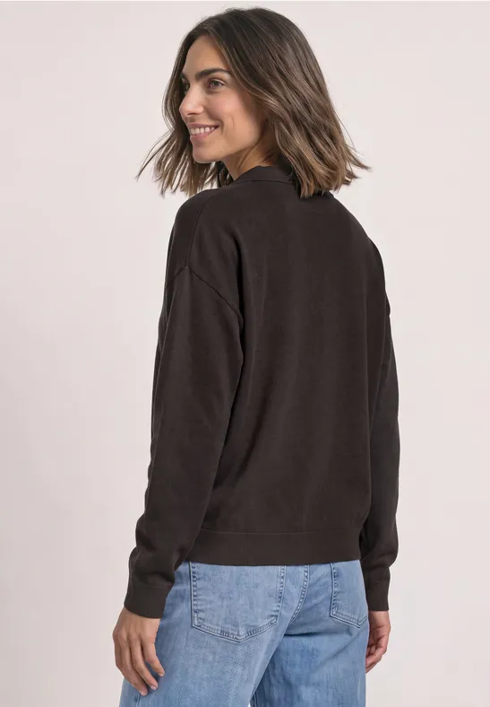 Femme en pull sombre et jean, regarde par-dessus l'épaule en souriant sur un fond clair.