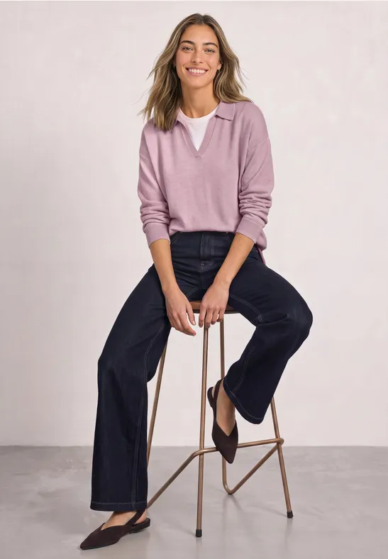 Femme souriante assise sur un tabouret, porte un pull rose col V et un jean large.