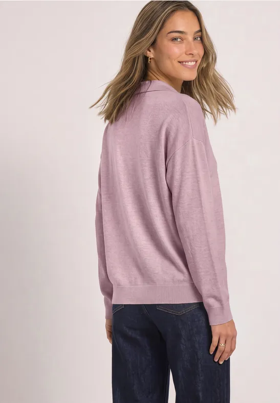 Femme en pull rose et jean foncé sourit par-dessus son épaule devant un fond blanc.