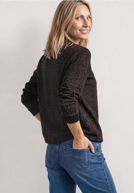 Frau in braunem Pullover und blauen Jeans, seitlich stehend mit Händen in den Taschen, lächelnd.