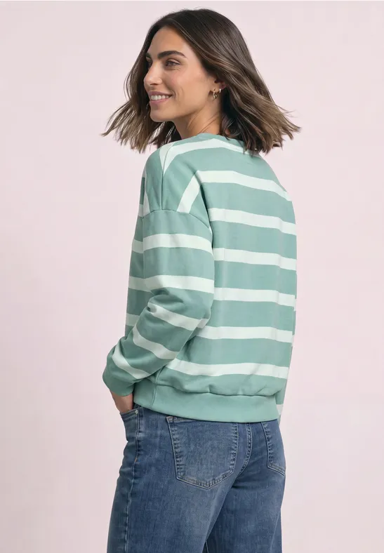 Frau im grün-weiß gestreiften Pullover, seitlich stehend, lächelnd.