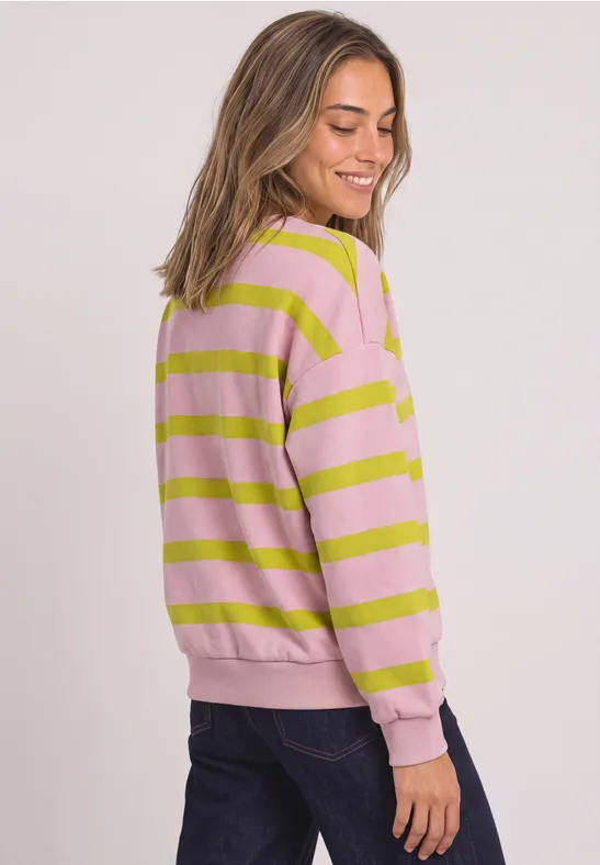 Frau im rosa-grün gestreiften Pullover und dunkler Jeans, seitlich stehend und lächelnd.