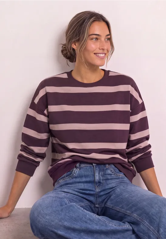 Frau in gestreiftem Pullover (rosa/lila) mit Jeans, sitzt lächelnd vor hellem Hintergrund.