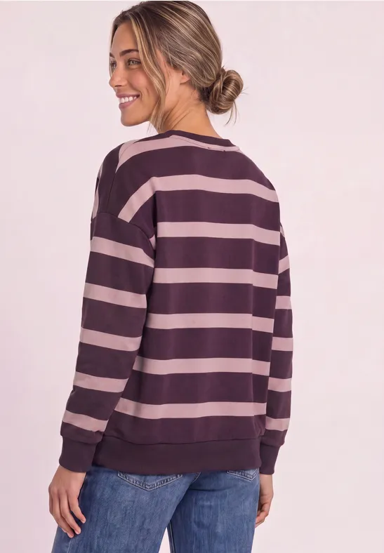 Frau mit locker sitzendem gestreiftem Pullover (lila und rosa) und Jeans, Gesicht lächelnd zur Seite gewandt.