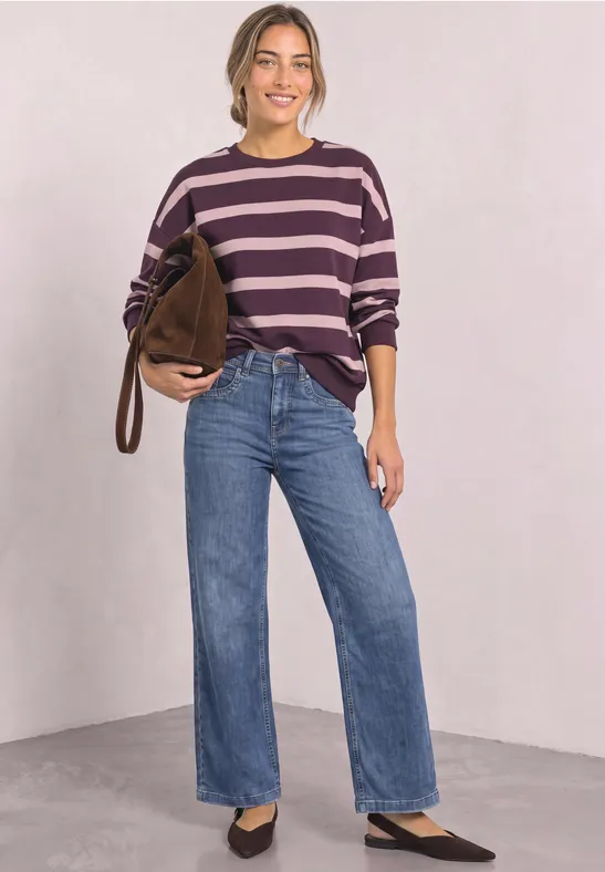 Frau in lila-gestreiftem Pullover, weiten blauen Jeans und schwarzen Schuhen, mit brauner Tasche vor heller Wand.