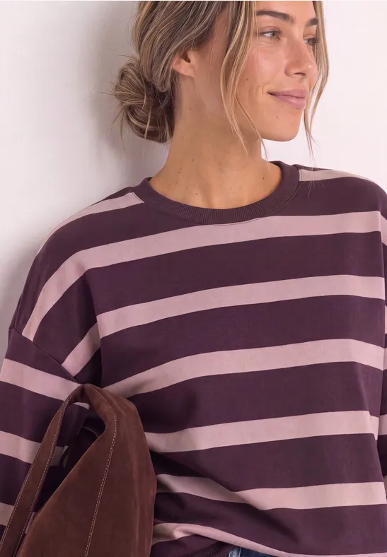Frau in gestreiftem Pullover (weinrot und rosa) mit brauner Tasche, seitlich lächelnd vor weißer Wand.