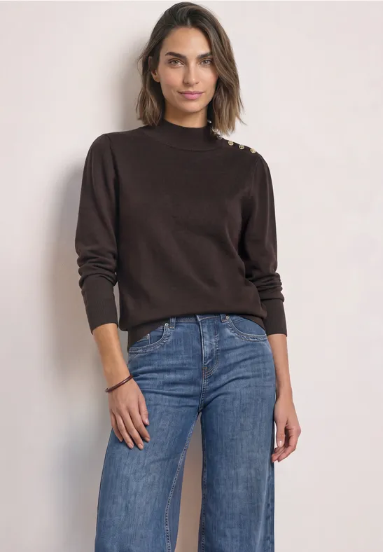 Vrouw met donkeren trui met schouderknopen en blauwe jeans voor een lichte achtergrond.