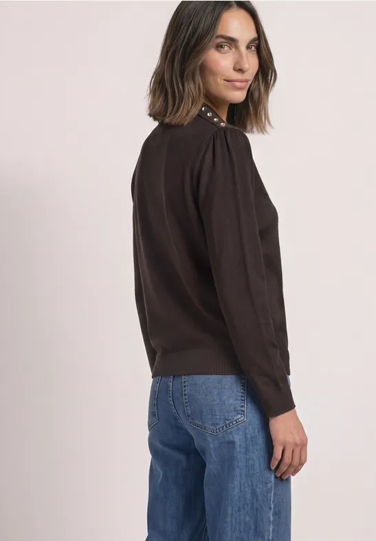 Vrouw in zwarte trui en blauwe jeans lacht terwijl ze over haar schouder kijkt.