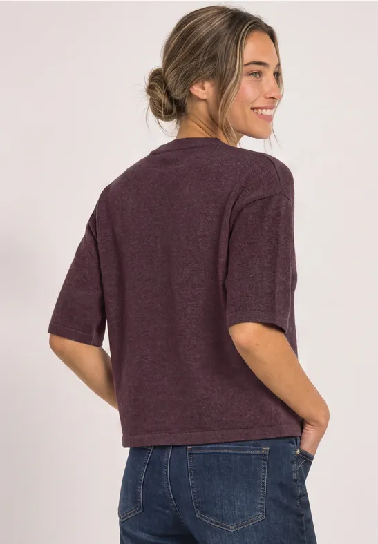 Frau mit Dutt im bordeauxfarbenen Kurzarm-Pullover und Jeans, seitlich lächelnd.