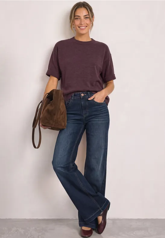 Frau in violettem T-Shirt, blauen Bootcut-Jeans und Loafers, trägt eine braune Wildledertasche.