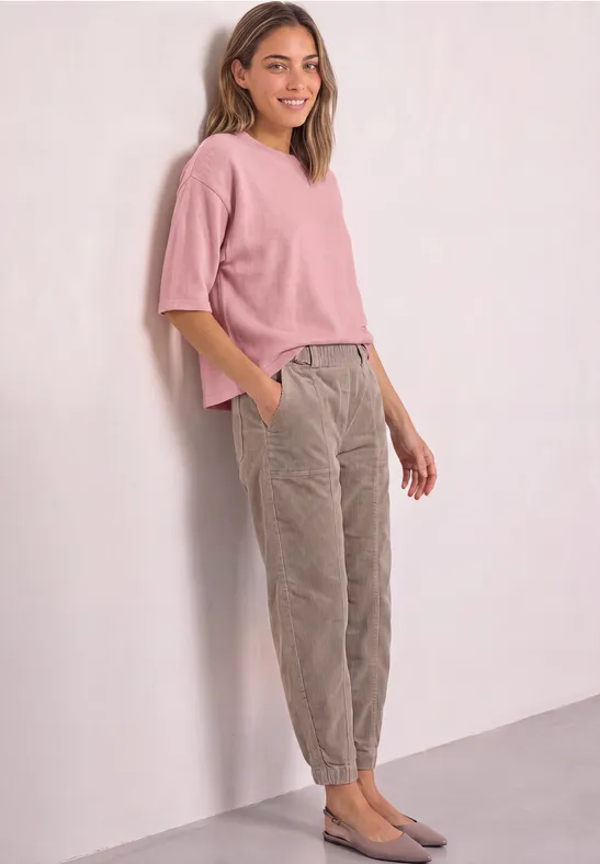 Eine Frau lehnt lächelnd an einer Wand, trägt ein rosa Oberteil, graue Cordhosen und graue Ballerinas.