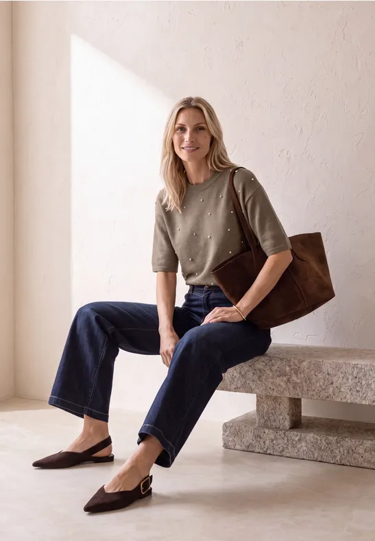 Blonde Frau in grünem Pullover, Jeans und brauner Tasche sitzt auf einer Bank in hellem Raum.