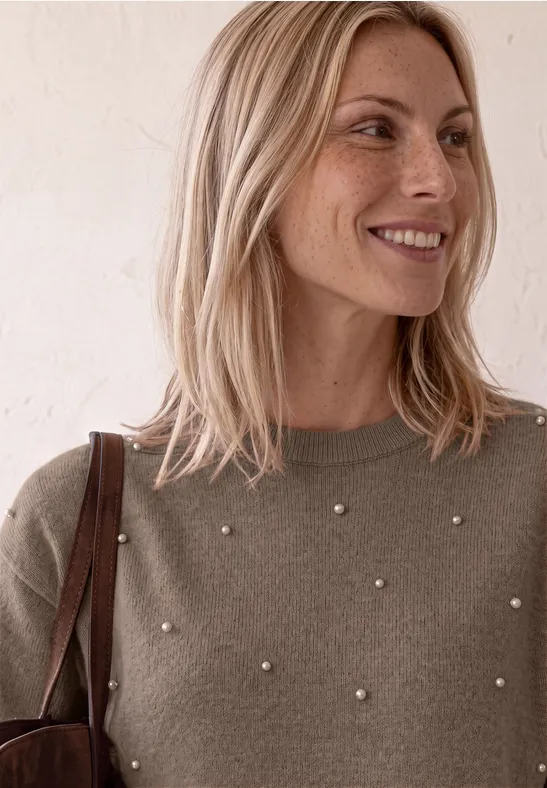 Eine blonde Frau mit Perlen-Verzierung am Pullover trägt eine Tasche und lächelt sanft seitlich.
