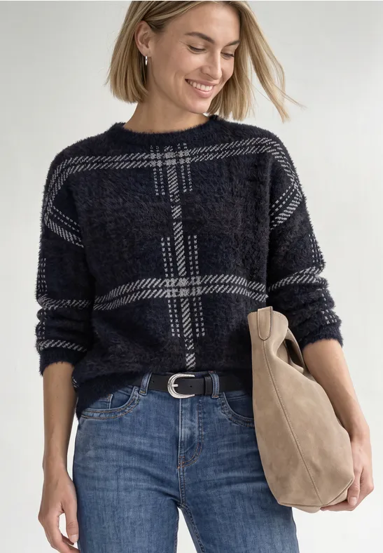 Een lachende vrouw met blond haar draagt een geruit trui, blauwe jeans en houdt een tas vast.