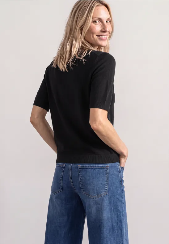 Eine Frau steht mit dem Rücken zur Kamera, schaut über die Schulter, trägt ein schwarzes Shirt und blaue Jeans.