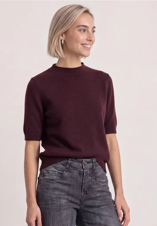 Eine junge Frau mit kurzem blondem Haar trägt einen dunklen Pullover und ausgewaschene Jeans.