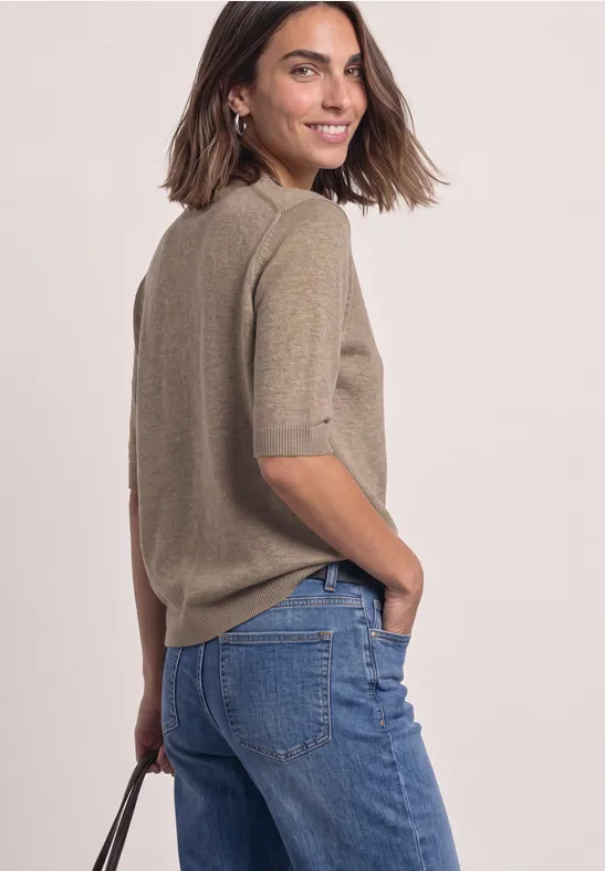 Frau in modischem, hellbraunem Kurzarm-Pullover und blauen Jeans, schaut über die Schulter und lächelt.