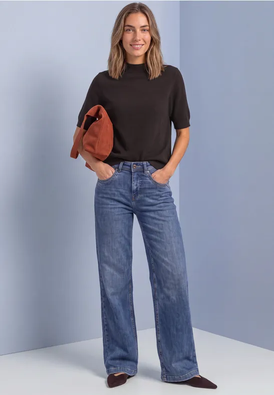 Frau im schwarzen T-Shirt und weiten Jeans trägt braune Jacke lässig über der Schulter vor hellblauem Hintergrund.