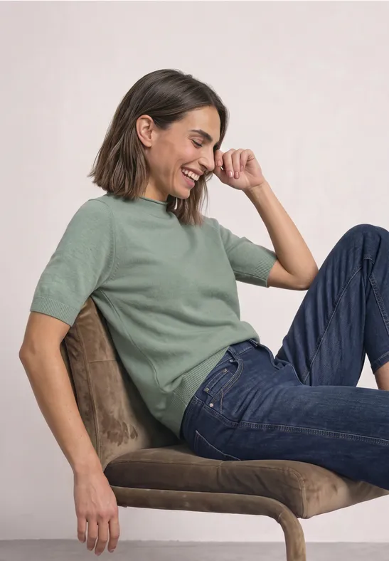 Frau im grünen Pullover und Jeans sitzt seitlich auf einem Stuhl und lacht.