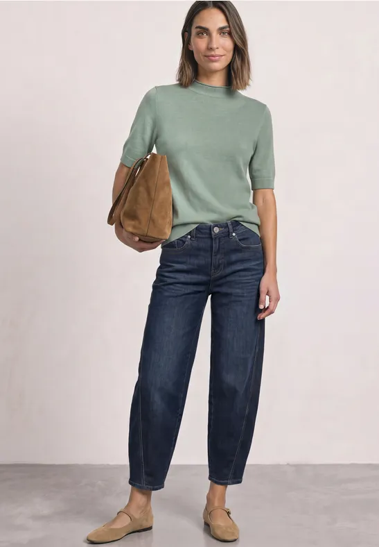 Frau mit olivgrünem Pullover, blauen Jeans, beigen Schuhen und brauner Tasche vor schlichtem Hintergrund.