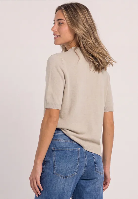 Frau in beigem Kurzarm-Pullover und blauen Jeans, seitlich mit Blick nach links vor hellem Hintergrund.