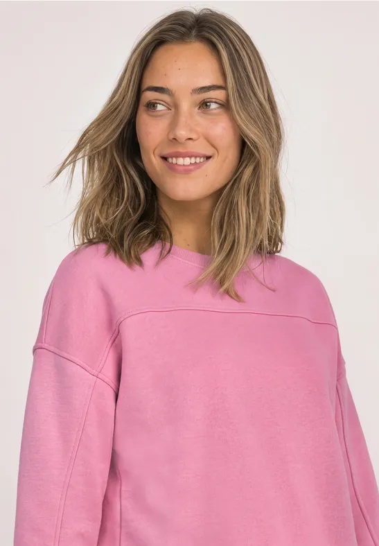 Eine junge Frau in rosa Sweatshirt lächelt vor hellem Hintergrund.