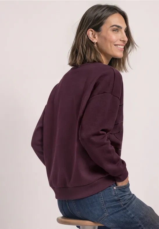 Eine lächelnde Frau im lila Pullover und Jeans, sitzt seitlich und blickt über die Schulter.