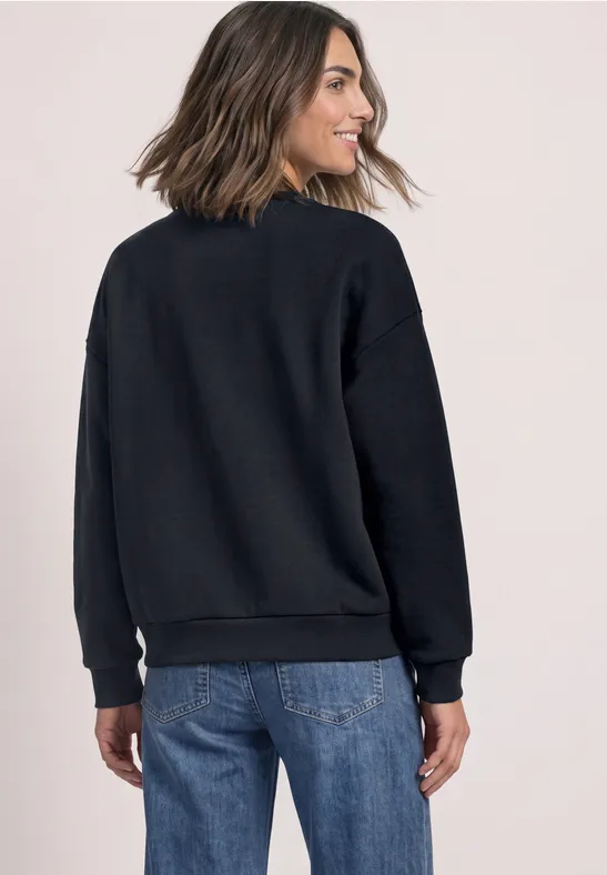 Frau trägt schwarzen Pullover und Jeans, schulterlanges Haar, sie ist halbseitig von hinten zu sehen.
