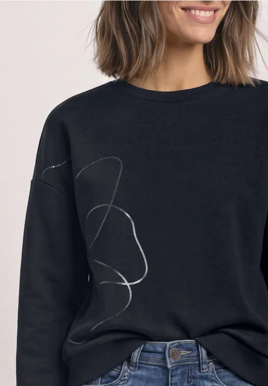 Frau trägt schwarzen Pullover mit abstrakt geschwungenem weißen Linienmuster.