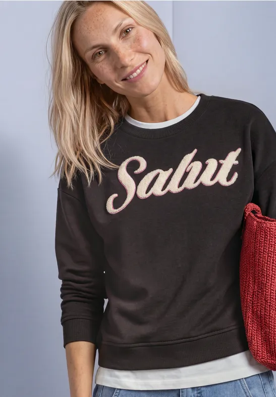 Uśmiechnięta blondynka ma na sobie czarną bluzę z napisem „Salut” i trzyma czerwoną torbę.