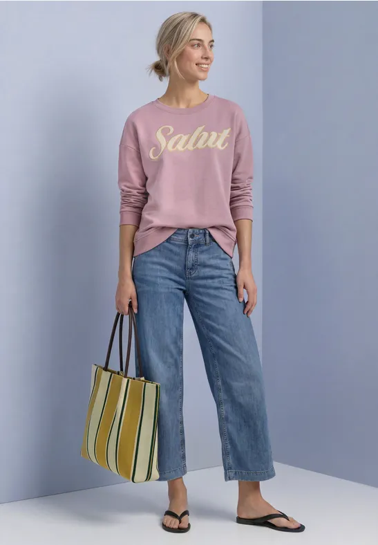 Frau in rosa Pullover mit Schriftzug "Salut", weiter Jeans, gestreifter Tasche und Sandalen.