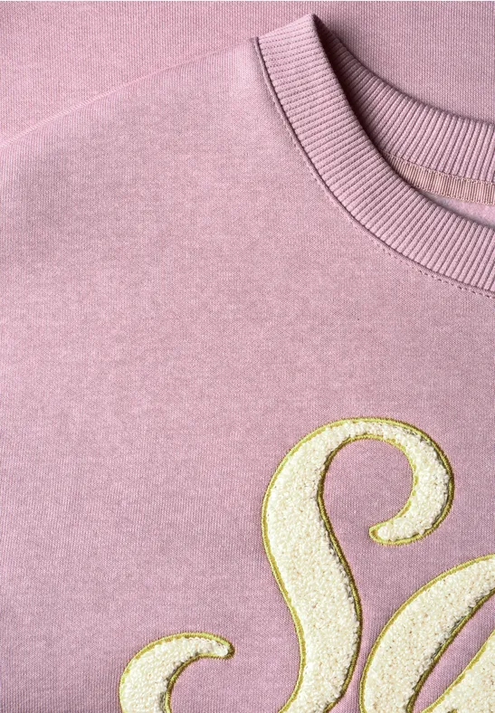 Rosafarbener Pullover mit goldglitzerndem Buchstaben-Detail, Nahaufnahme.
