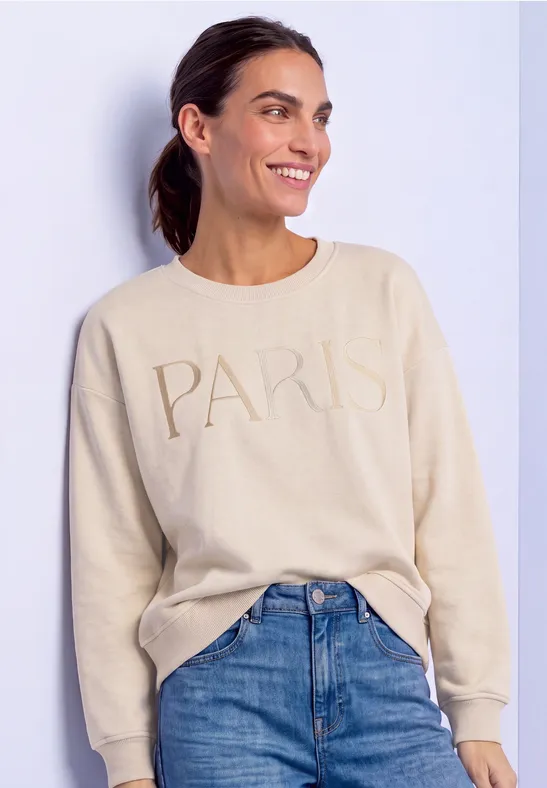 Frau in beigem Pullover mit 'PARIS'-Schriftzug und Jeans lächelt seitlich.