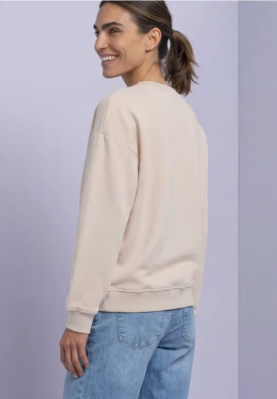 Junge Frau im beigen Pullover und Jeans, seitlich stehend vor lila Hintergrund, lächelnd.
