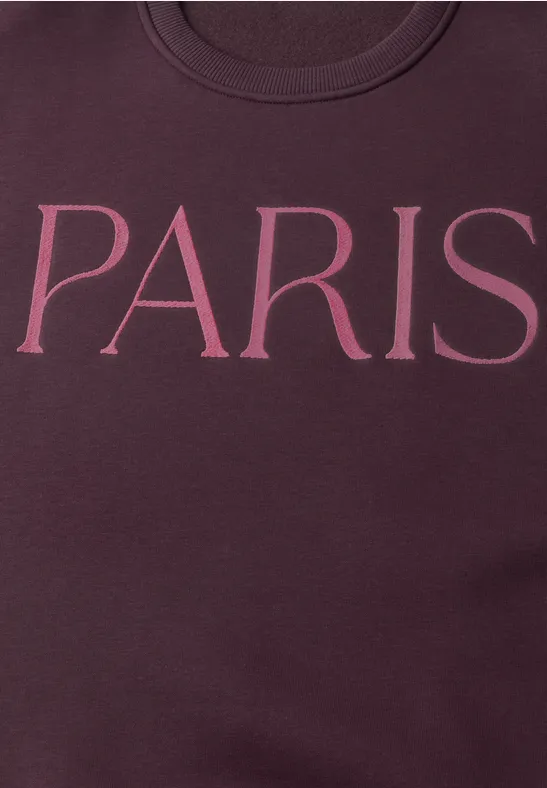 Close-up van een bordeauxkleurige sweater met de tekst 'PARIS' in grote roze letters.